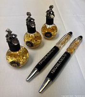 Three decorative glass bottles with suspended gold leaf flakes, each topped with silver-tone miniature gold miner figurine caps, alongside two Romarco Minerals branded pens with gold leaf flakes inside the upper transparent chamber.