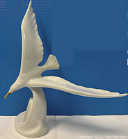 Glossy white porcelain sculpture of a seagull in flight with detailed golden beak and smooth curved base, placed against blue background.