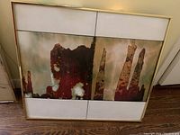 Framed abstract enamel artwork with lines dividing into four panels, earthy rust and white colors, gold-tone frame shown leaning on floor.