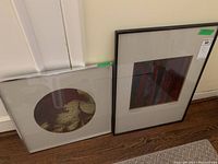 Two framed art prints leaning against the wall on a wooden floor, showing their full size and framing.