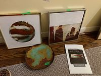 Two framed enamel artworks leaning against wall on floor, enamel bowl and booklet in front.