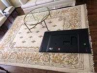 Rectangular cream colored area rug with decorative tan and beige floral and geometric pattern under glass coffee table and beside sofa and chairs. Rug has fringe edges and visible wear.