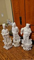 Three white porcelain wise man figurines on wooden floor in front of a cabinet, each standing on cloud-shaped bases