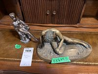 Two brass figures on a wooden surface in front of cabinetry. Larger reclining male figure, smaller standing female figure. Green tags with dimensions and lot label visible.