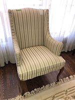 Front angle view of a wingback chair with beige and green vertical stripes on fabric upholstery, wooden legs, placed on hardwood floor near sheer curtains.