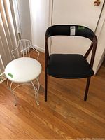Photo showing two chairs side by side on wooden floor; one white metal frame with round vinyl seat, one dark wood frame with black faux leather seat.