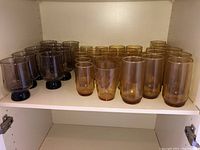 Full set of 24 assorted glasses arranged on a cabinet shelf showing different colors and styles.