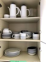 Multiple shelves showing organized white Meakin ceramic dishware with black geometric rim pattern including coffee pots, plates, creamers, sugar containers, and bowls.
