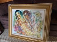 Framed painting displayed on floor against a sofa showing two nude female figures in an abstract style, gold frame and white matting visible.
