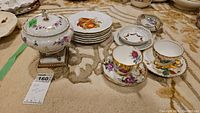 Wide shot showing lidded pedestal bowl, stacked fruit plates, and two floral English teacups with saucers on a carpeted surface.