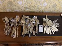 Full view of assorted silverplate and plastic-handled utensils laid out on a wooden surface including knives, forks, spoons, and small shakers.