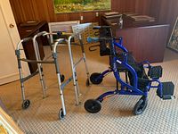 Photo showing two standard walkers and the Fusion wheelchair walker together in a room with wood paneling and carpeted floor.