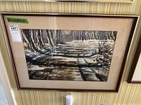 Framed watercolour painting depicting a winter woodland path with shadows and snow, shown hung on striped wallpapered wall.