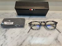Prescription eyeglasses with marbled grey acetate frame, silver-tone arms with red-white-blue tips, paired with black textured case and branded cleaning cloth displayed on marble surface
