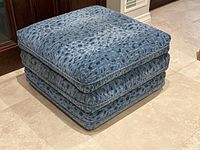 Front-left angle of ottoman on tiled floor showing upholstery pattern and piped trim