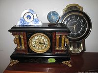 Photo showing four vintage clocks on a wooden surface, prominent is a large black and gold mantel clock, behind it a large round wall clock with silver trim, and two smaller blue and white clocks on top.