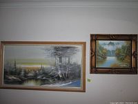 Two framed paintings side by side. Left is a wooden frame with river and trees; right is an ornate golden frame with mountain and lake landscape.