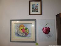 Three framed art prints hung on wall: floral above, horn of plenty with fruit left, and apple/cherry painting right