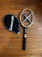Top-down view of the wooden tennis racket and its cover placed on a wooden floor. The cover is black with white Bancroft branding. The racket is natural wood with a black handle and metal cross braces on the throat.