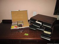 Overall view of silver cassette tape storage case partially filled with tapes and two wooden slide-out storage boxes with drawers for tapes, on wooden table.