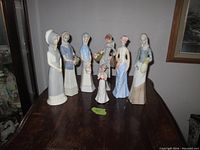 Eight ceramic figurines of women in traditional and vintage attire displayed on a dark wood table.