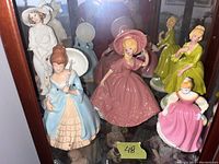 Group of seven ceramic figurines featuring women in period dresses of varying colors and styles, displayed inside a glass cabinet.