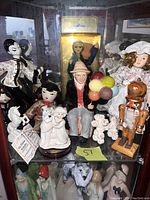 A collection of assorted decorative figurines including porcelain dolls, balloon man figurine, crocheted poodle, nutcracker, and small animal figures displayed together in a cabinet.