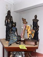 Six figurines on a wooden surface including three dark bronze African-style figurines, a pink dress porcelain lady with child and a carousel horse figurine