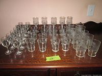 Front angle viewing a large assortment of clear glassware including shot glasses and stemmed glasses arranged on a wooden surface.