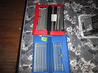Four sets of metal knitting needles arranged in two storage cases, one red and one blue, showcasing various sizes.
