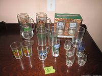 Photo showing assortment of novelty glasses and mugs arranged on surface including boxed set of shot glasses.