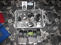Top-down view showing five Panasonic cordless phone handsets arranged in a row with numeric keypads and display screens visible. Wired charging bases and cords placed in plastic tray alongside phones.