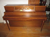 Photo of upright brown wooden piano with music stand and three brass pedals from the front.