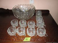 Set shows one large decorative glass punchbowl with a scalloped edge and a textured diamond pattern and eleven glass cups with handles in front, each cup featuring the same pattern.