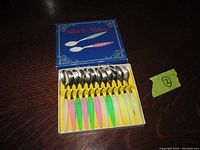 Photo of the set of 12 tiny spoons arranged in yellow insert inside the blue box, showing spoon heads and pastel colored handles.