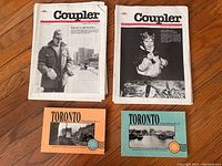 Seven Coupler magazine issues stacked with two Toronto postcard books in front, placed on wooden surface. Covers show historical black and white photos.