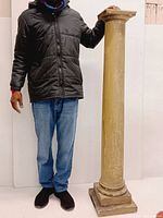 Photo showing the full height of the architectural wood column with a person standing next to it for scale. The column has a square base and a decorative top and is painted in a faded beige color with visible weathering.