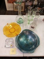 Top view of wine chiller decanter, green-tinted goblets, brandy decanter, amber glass bowl, blue-green art glass bowl, and square crystal candle holder on white table.