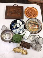 Overview of the lot showing collector's plates, green enamelware teapot, metal car plaque, metal coasters, and decorative plaques on white table