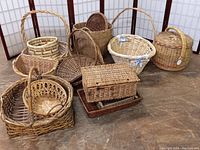 View of ten hand made woven baskets arranged on floor in front of folding screen. Baskets include round, oval, rectangular shapes in various natural fiber colors and styles.