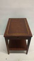 Front view of rectangular wooden end table showing leather top with gold border, drawer knob, and lower shelf.