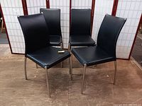 Four modern style dining chairs with chrome metal frames and black vinyl padded backs and seats arranged on a floor in front of a folding room divider.