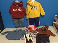 Various boys' clothing items for 12-24 months displayed on a blue background, showing the full lot including hoodies, pants, button-up shirts, and a plush dog toy.