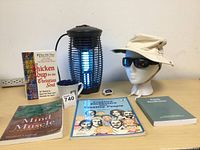 Wide shot of all items on table including bug zapper, books, mug, pedometer, hat with sunglasses on mannequin head.