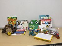 Photo shows a collection of assorted children's toys and learning materials including a green owl backpack, a brown owl plush puppet, children's books, Crayola crayons, a yellow pegboard with pegs, a yellow drawing board, toy dinosaur, CDs, and small plush ball and hat toys.