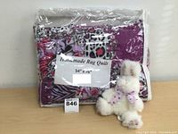 Photo of the handmade rag quilt packed in plastic bag with a label showing dimensions 54x76, alongside the white stuffed bunny rabbit with purple bow.