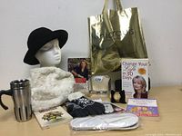 Photo showing black hat on mannequin, white scarf, stainless steel coffee mug, two books, gloves, slippers, wristwatch in box, gold Marquis bag, and a mannequin showcasing scarf and sunglasses.
