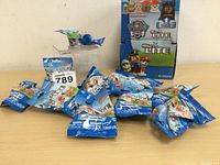 Dozen Paw Patrol Micro Lites in sealed blue packaging scattered in front of an open Paw Patrol Micro Lite box showing cartoon character artwork.