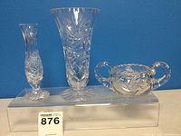 Three crystal pieces on display against blue background: small slender vase, tall flared vase, double-handled bowl, all with pinwheel patterns