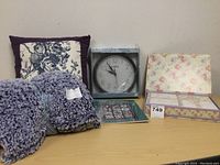 Photo showing pillow, quilt, wall clock in box, letter paper with envelopes, and Quilters Almanac book on table.